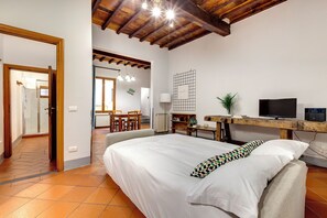 Relax in this inviting room with a large bed, desk, and TV, surrounded by a charming wooden beam ceiling and a small potted plant.
