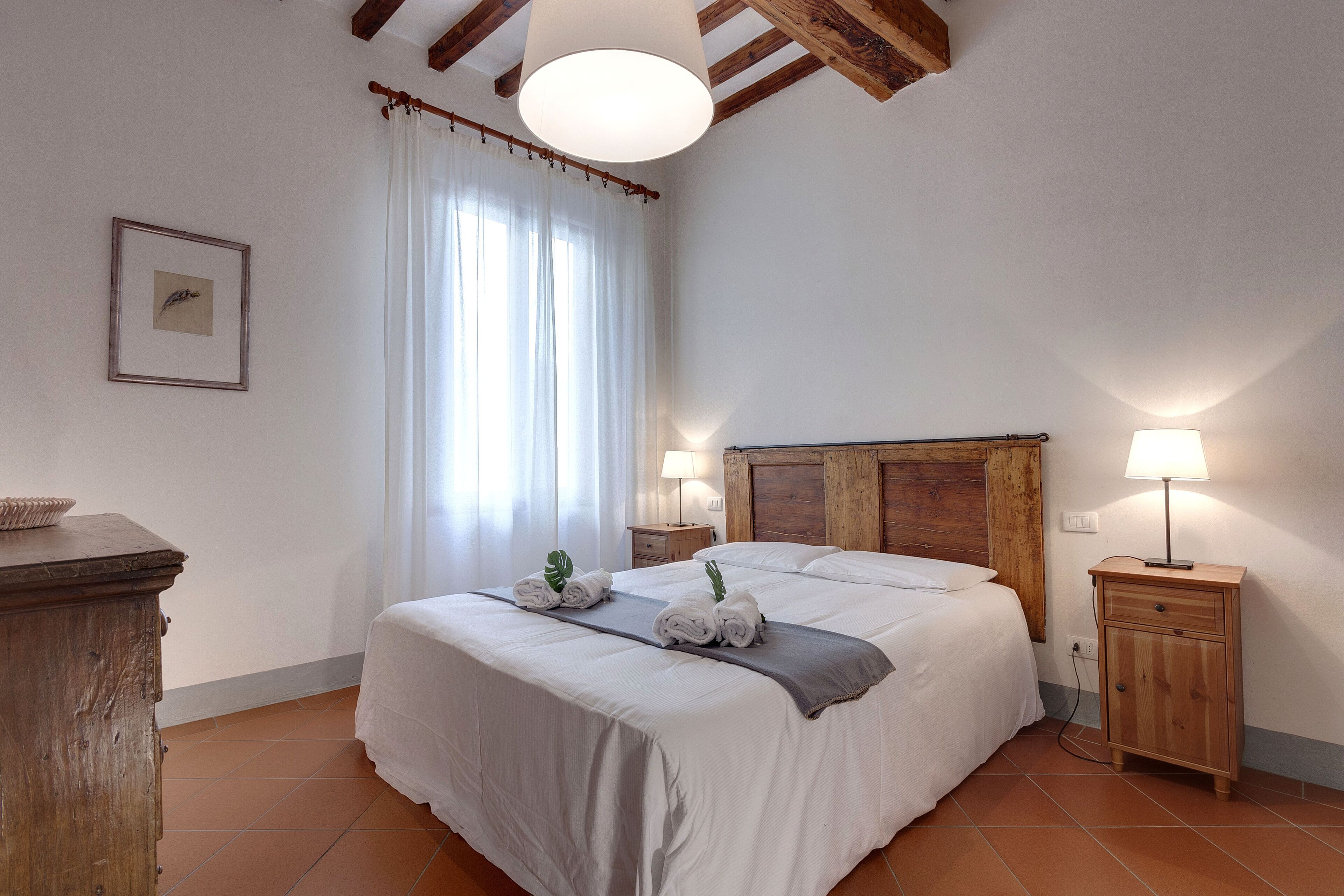 A neatly made bed with towels, a bedside lamp, and a wooden nightstand in a room with white walls and a wooden ceiling beam.