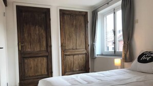 A bedroom with a bed, two wooden doors, a window with curtains, and a wall-mounted lamp. - Authentic house (6p) in prime location near Maastricht (Eijsden)