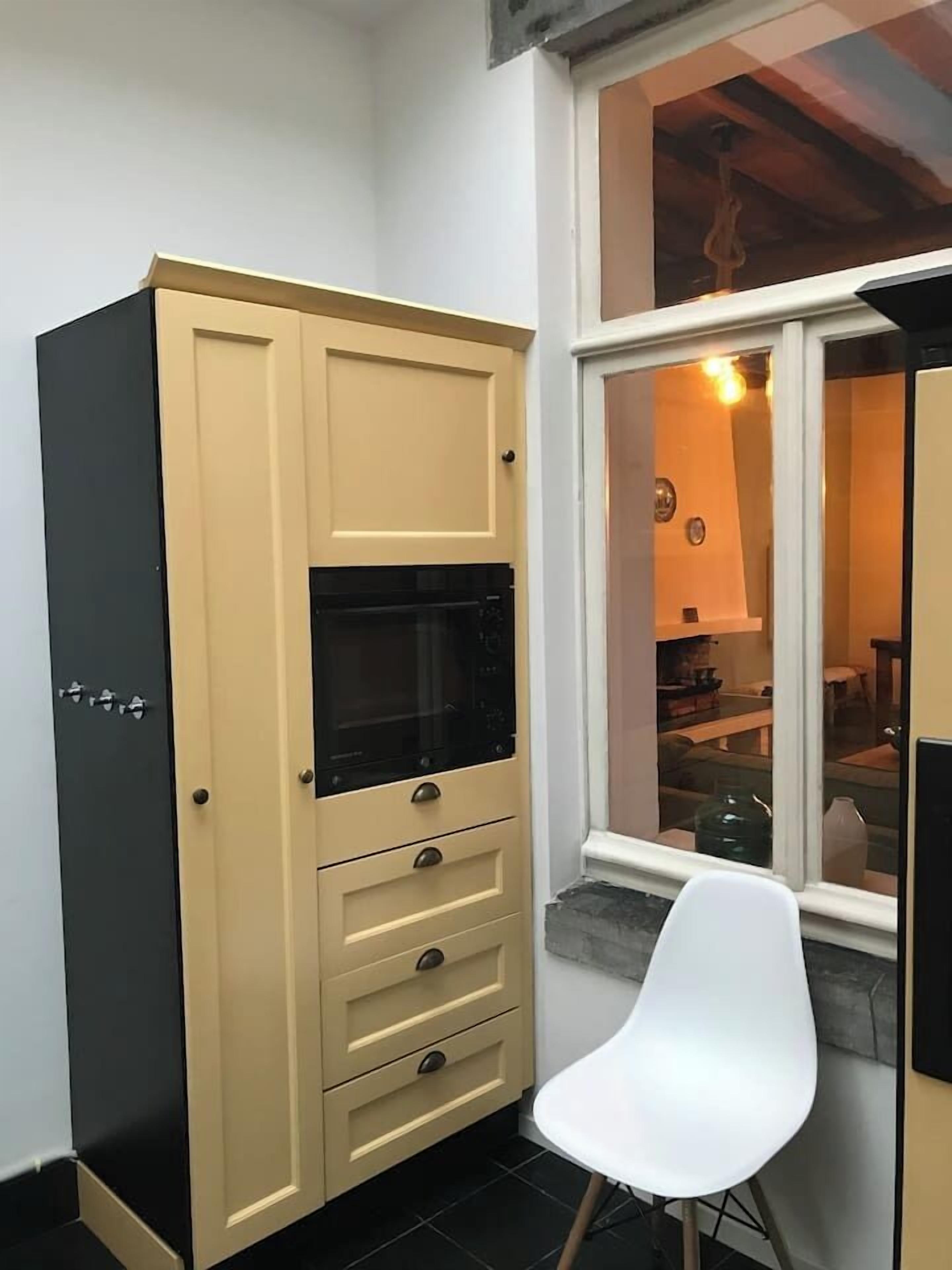 Eine Küchenzeile mit Mikrowelle, einem Schrank mit Schubladen, einem weißen Stuhl und einem Fenster mit Blick ins Innere.
