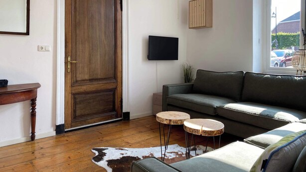 Un salón con puerta de madera, sofá gris, dos mesas redondas de madera, televisor montado y ventana con vistas a coches estacionados.