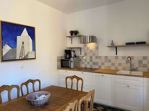 Une cuisine compacte avec des meubles blancs, un four à micro-ondes, une machine à café et un évier. Un tableau est accroché au mur, et une table à manger avec des chaises se trouve dans la pièce.