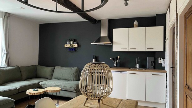 Un salon moderne comprenant un canapé, une table à manger décorée de bougies, un luminaire suspendu original et un coin cuisine avec des placards et un évier.