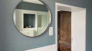 Une salle de bain avec un miroir rond, des carreaux blancs, une porte en bois et un interrupteur.