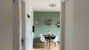A hallway leading to a dining area with a table and chairs, a pendant light, and a wall-mounted shelf with decorative items. - Cozy apartment (4p) prime location near Maastricht (Eijsden)