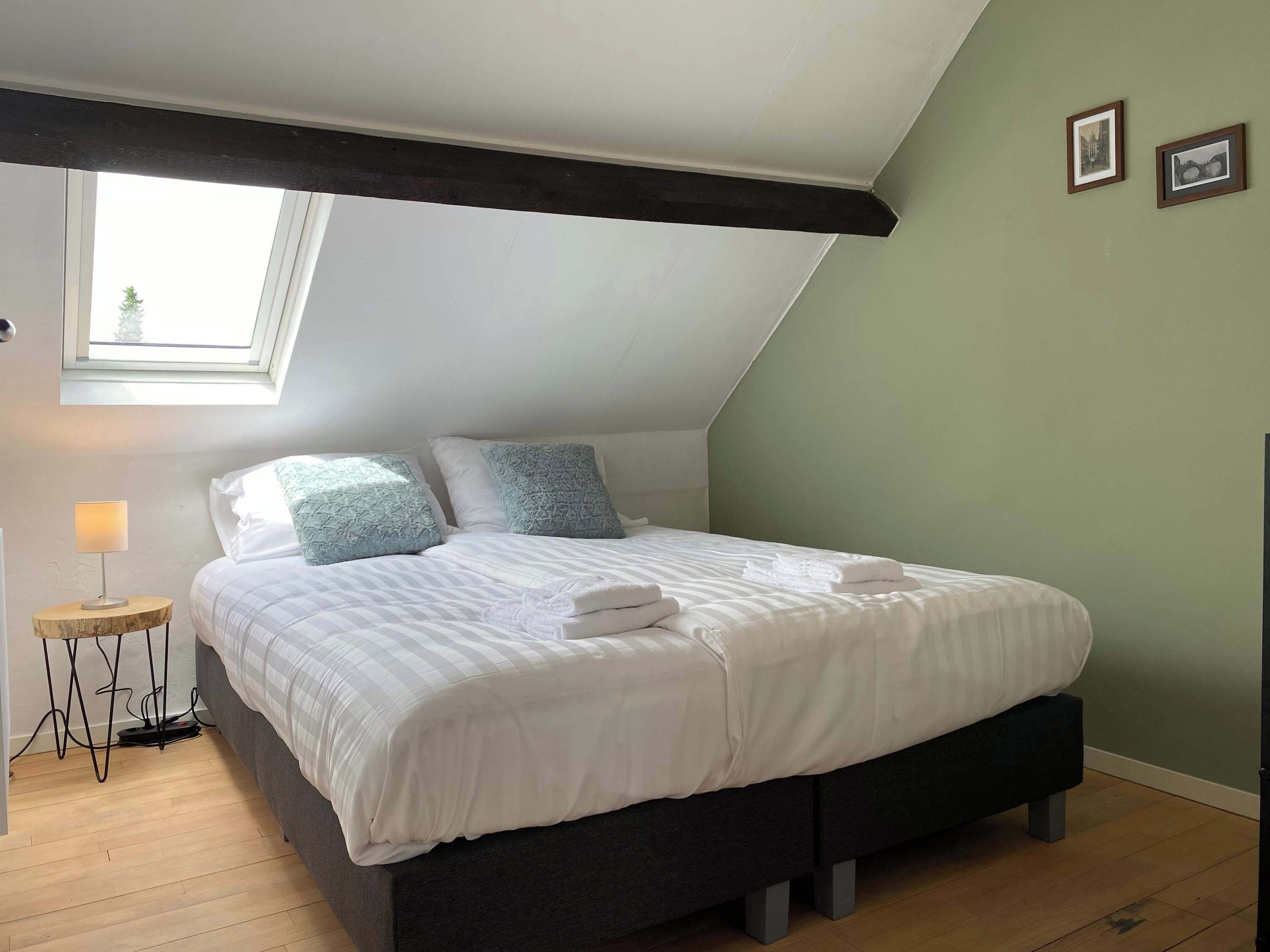 Een slaapkamer met een schuine kap, dakraam, een bed met wit beddengoed, twee kussens, een nachtkastje met een lamp, en twee ingelijste foto’s aan de muur.