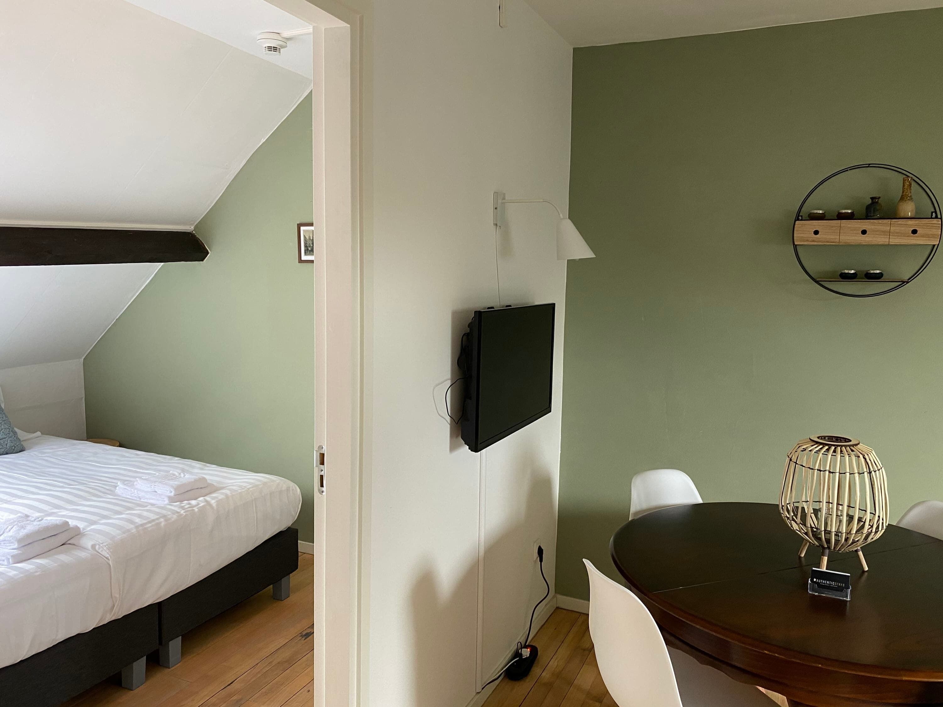 Ein Zimmer mit einem Bett, einem an der Wand befestigten Fernseher, einem Esstisch mit Stühlen und einer kleinen Ablage mit Dekoration.