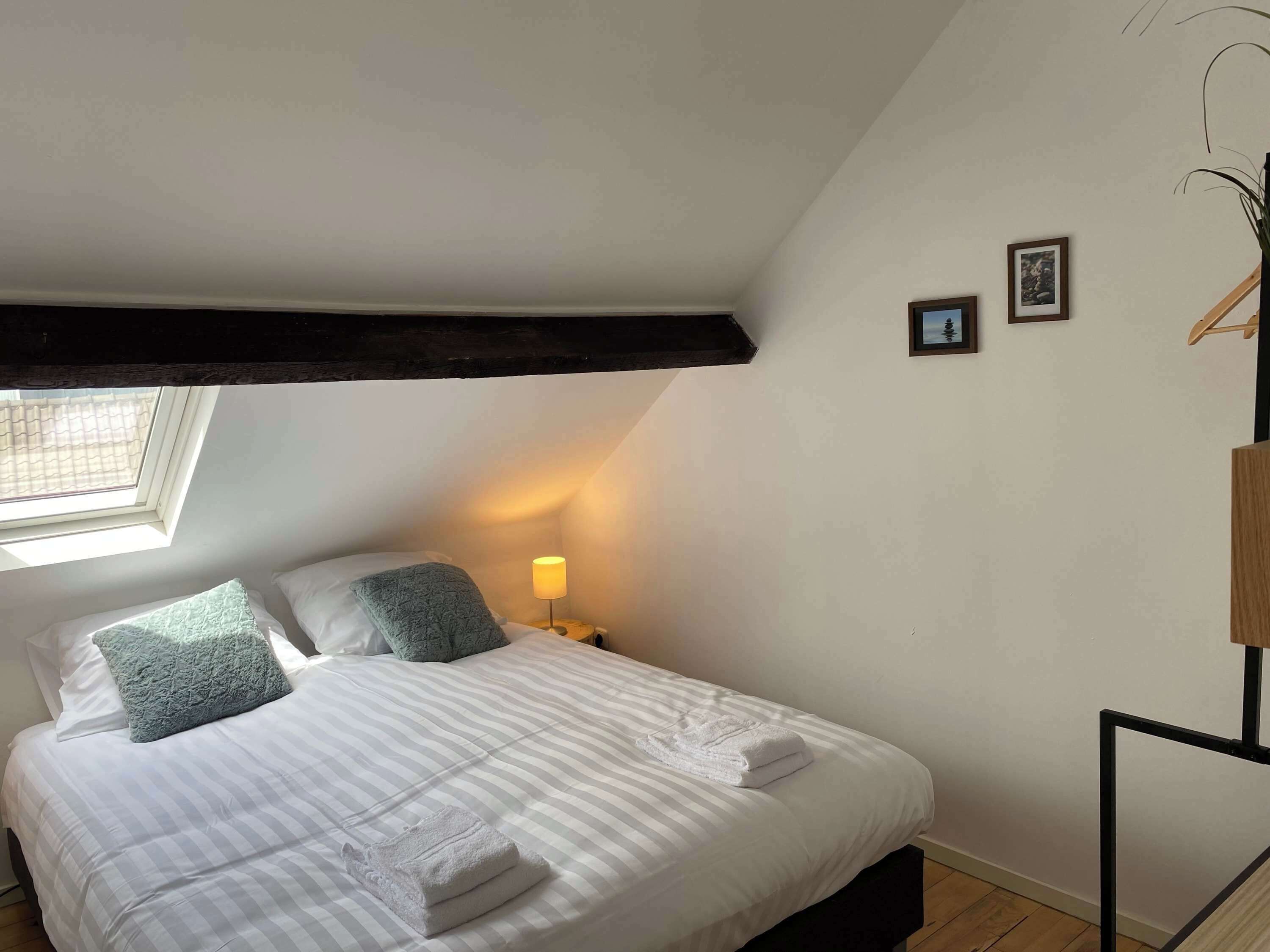 Een slaapkamer met een bed, kussens, een bedlampje, een raam en een klein schilderij aan de muur.