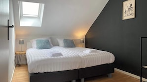 A bedroom with a bed, bedside lamps, a small table, a window, and a dark wall with a picture. - Atmospheric apartment (5p) in toplocation near Maastricht (Eijsden)