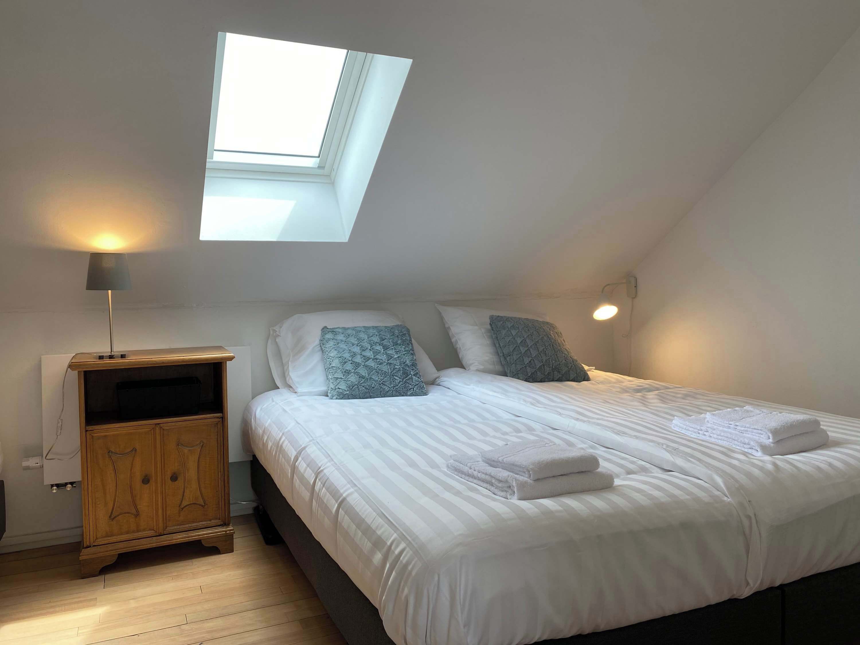 Een slaapkamer met dakraam, nachtkastje met lamp, houten kast, bed met wit beddengoed en kussens, en een wandlamp.