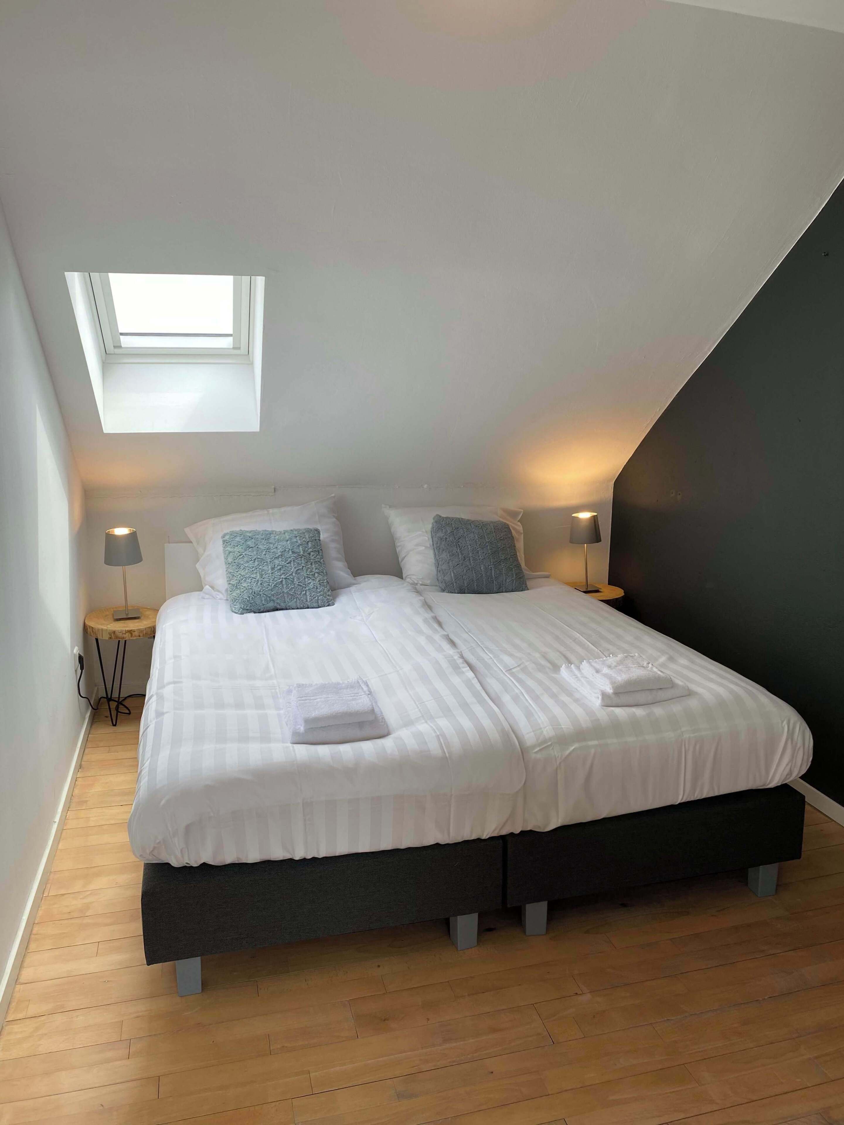 Un dormitorio con tragaluces, dos camas con ropa de cama blanca, almohadas grises, lámparas de noche y suelo de madera.