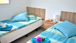 Ein Hotelzimmer mit zwei Einzelbetten, jeweils mit gestreifter Bettwäsche und einem Kopfkissen. Es gibt einen Nachttisch mit einer Lampe und einen kleinen Tisch mit Tasse und Untertasse.
