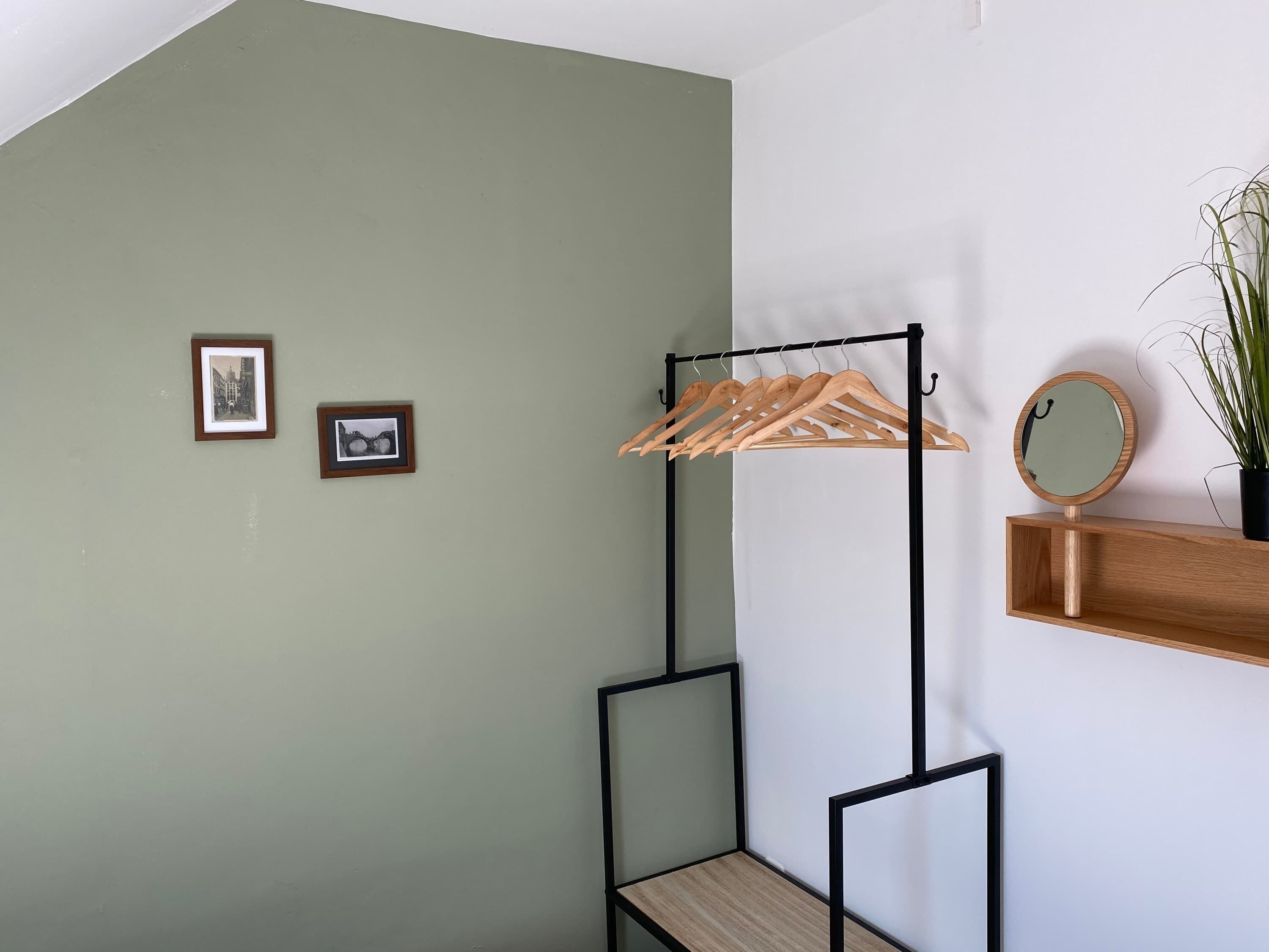 Una habitación con pared verde, perchero de madera, una pequeña estantería con un espejo y dos cuadros enmarcados.