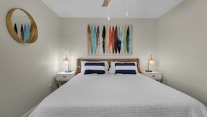 A bedroom with a large bed, two bedside tables, a ceiling fan, a wall-mounted mirror, and a colorful abstract painting. - Gulf Front Condo, Modern Coastal Vibe, Sleeps 6 + Free Attraction Tickets! (Santa Rosa Beach)
