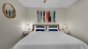 A bedroom with a large bed, two bedside tables, a ceiling fan, a wall-mounted mirror, and a colorful abstract painting.