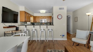 A modern kitchen with wooden cabinets, a microwave, and a built-in oven. There are white bar stools and a dining table with chairs. A flat-screen TV is mounted on the wall. - Gulf Front Condo, Modern Coastal Vibe, Sleeps 6 + Free Attraction Tickets! (Santa Rosa Beach)