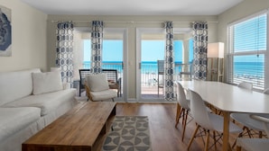 A living room with a white sofa, a wooden coffee table, and a dining area with a white table and chairs. There are patterned curtains and a view of the ocean through the glass doors. - Gulf Front Condo, Modern Coastal Vibe, Sleeps 6 + Free Attraction Tickets! (Santa Rosa Beach)
