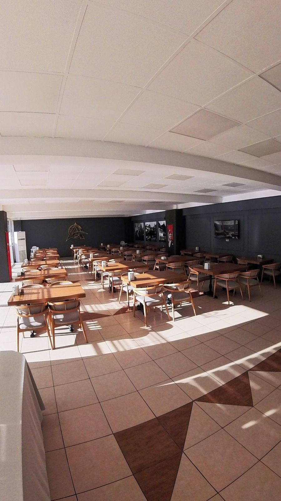 A spacious room with multiple wooden tables and chairs arranged in rows, a tiled floor, and a dark-colored wall with a mounted television.
