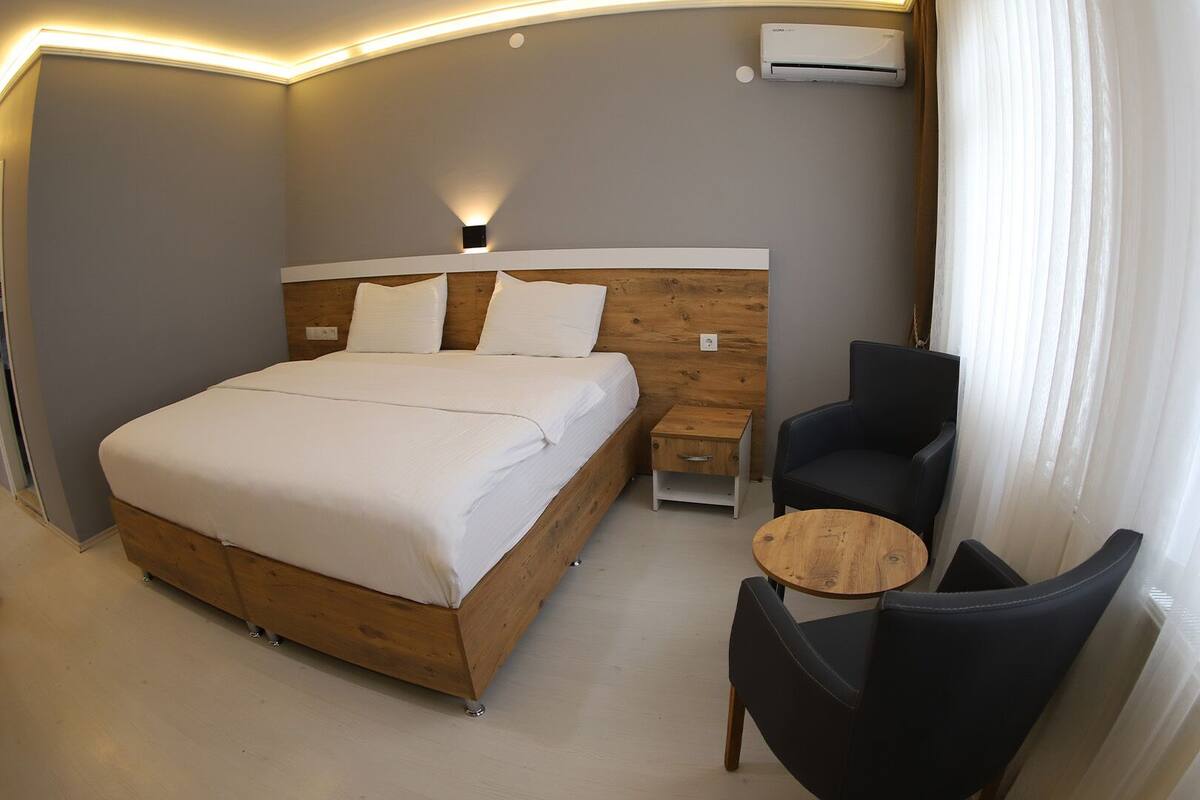 Una habitación de hotel moderna con una cama de madera, una mesita auxiliar pequeña, una mesa redonda de madera y una silla negra.
