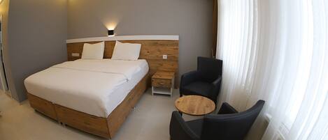 A modern hotel room with a wooden bed, a small side table, a round wooden table, and a black chair.
