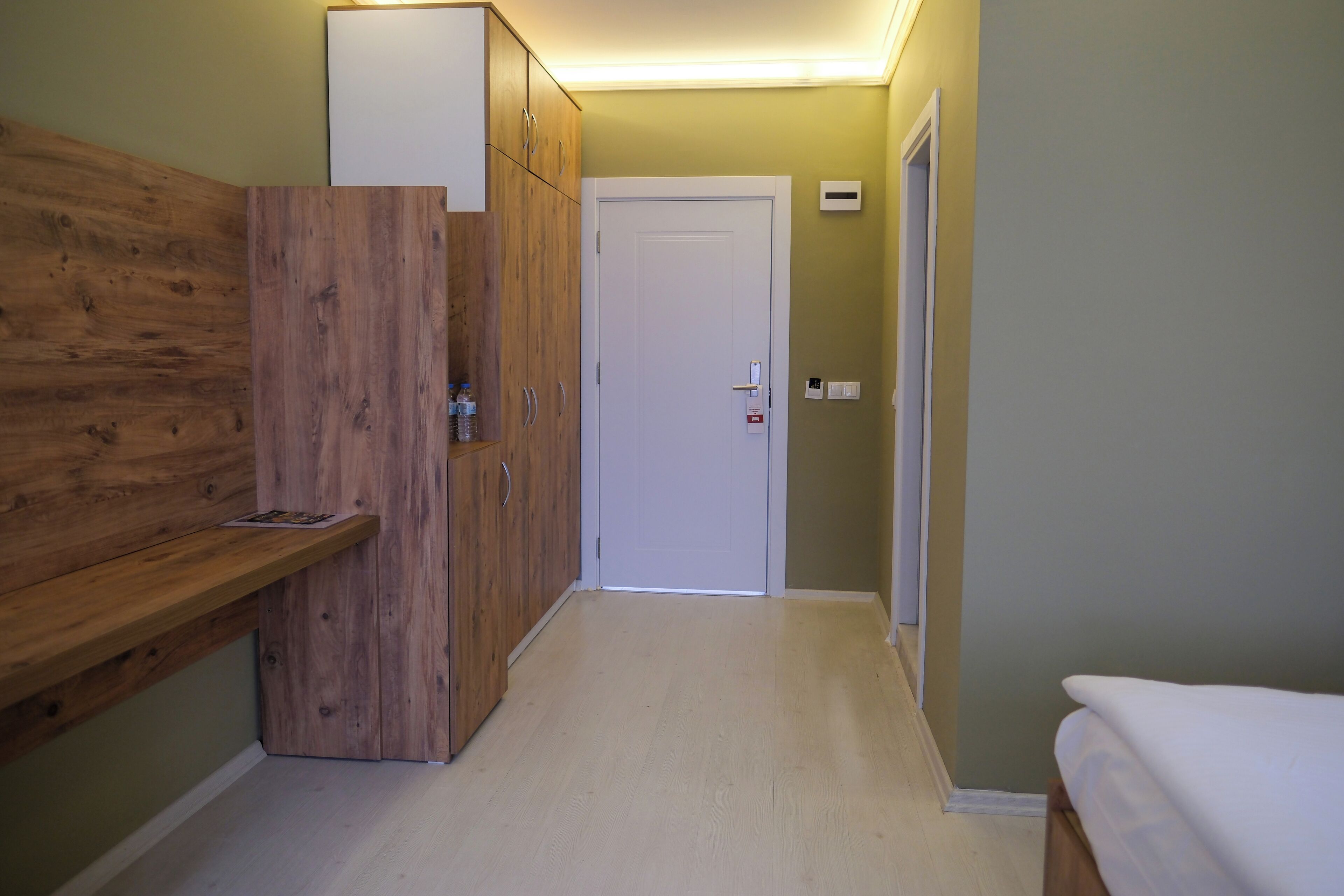A narrow hallway with wooden cabinets, a white door, and a bed with a white cover.