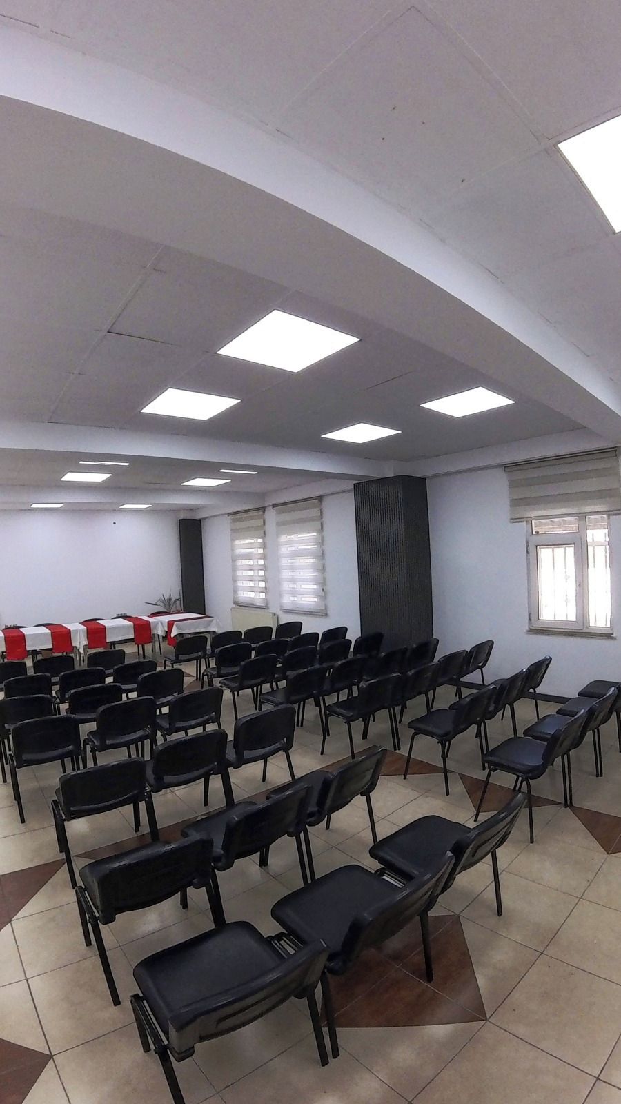 A conference room with rows of black chairs, white tables with red tablecloths, and large windows with blinds.