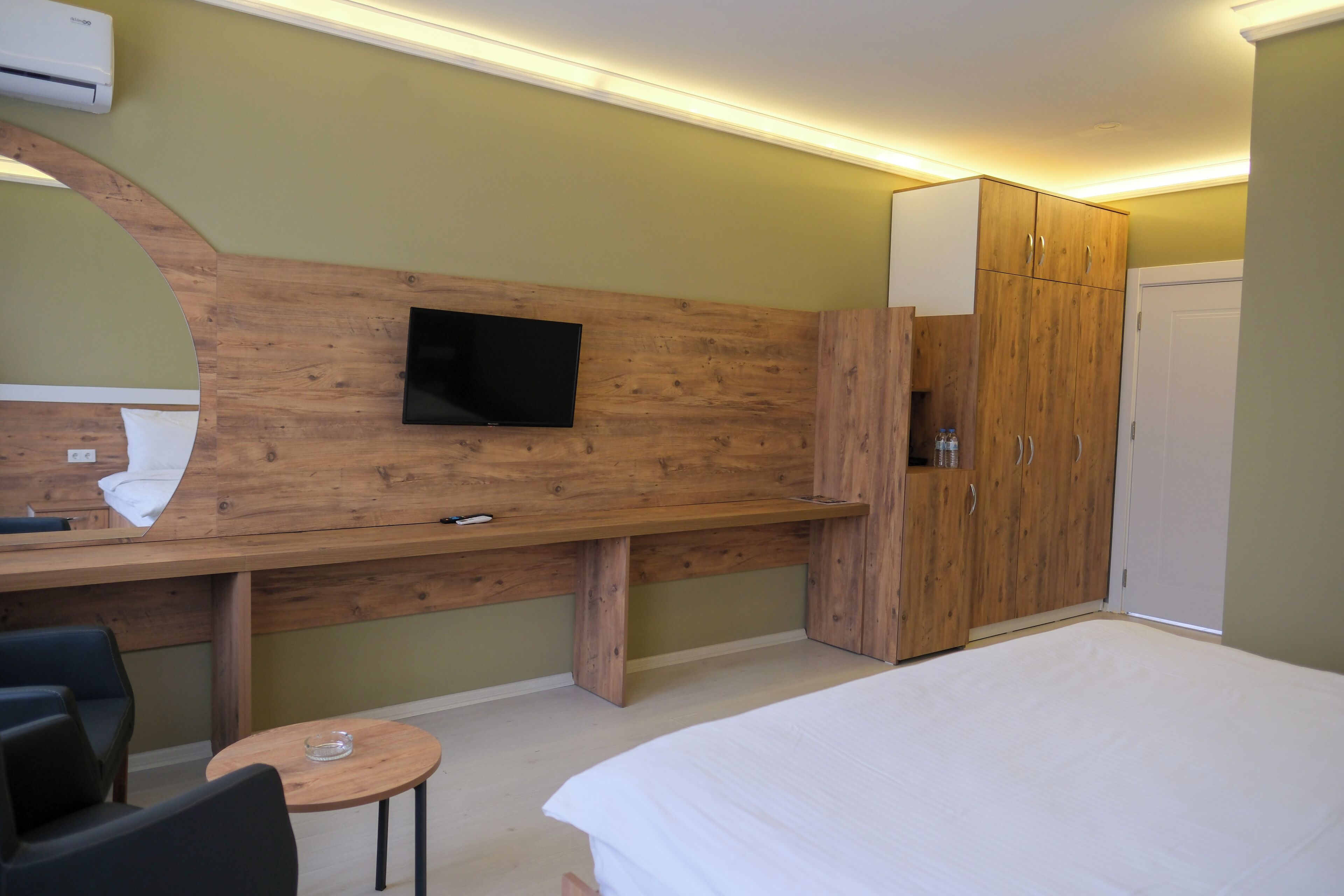 A hotel room with a wooden desk, a television mounted on the wall, a wardrobe, a bed, and a small round table.
