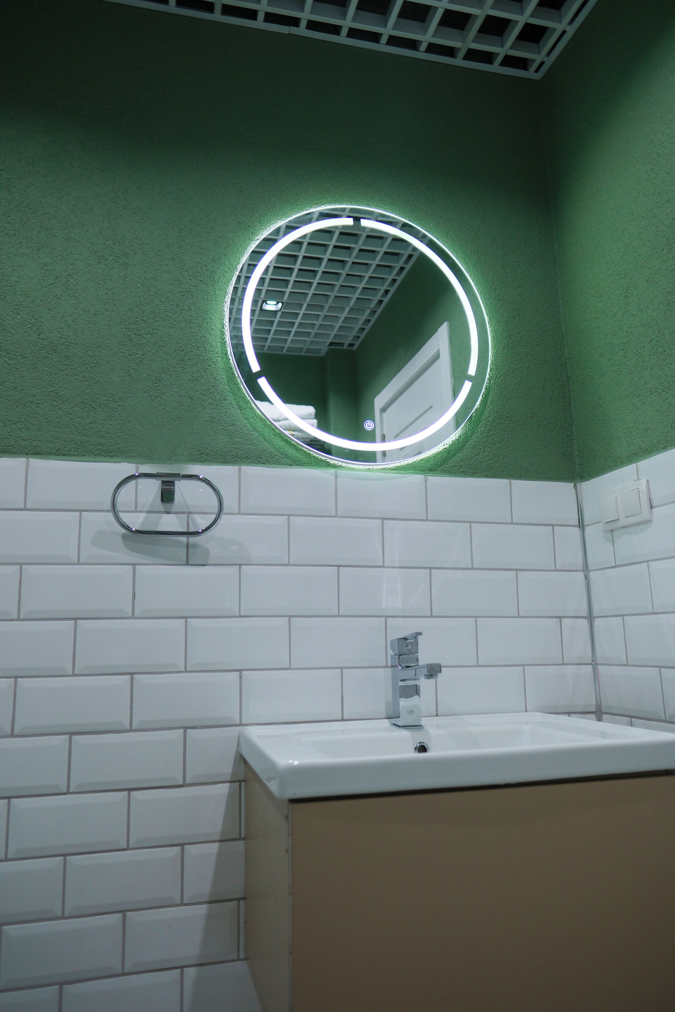 A bathroom with a white sink, green walls, and a circular mirror with a light.