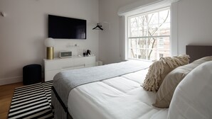 A modern bedroom with a large bed, a flat-screen TV mounted on the wall, a bedside table with a lamp, and a window with a view of buildings.