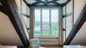 A room with a high ceiling, exposed wooden beams, a large window with a view of buildings, a chair, and a radiator. - Hitrental Chapel Bridge Apartments (Lucerne)