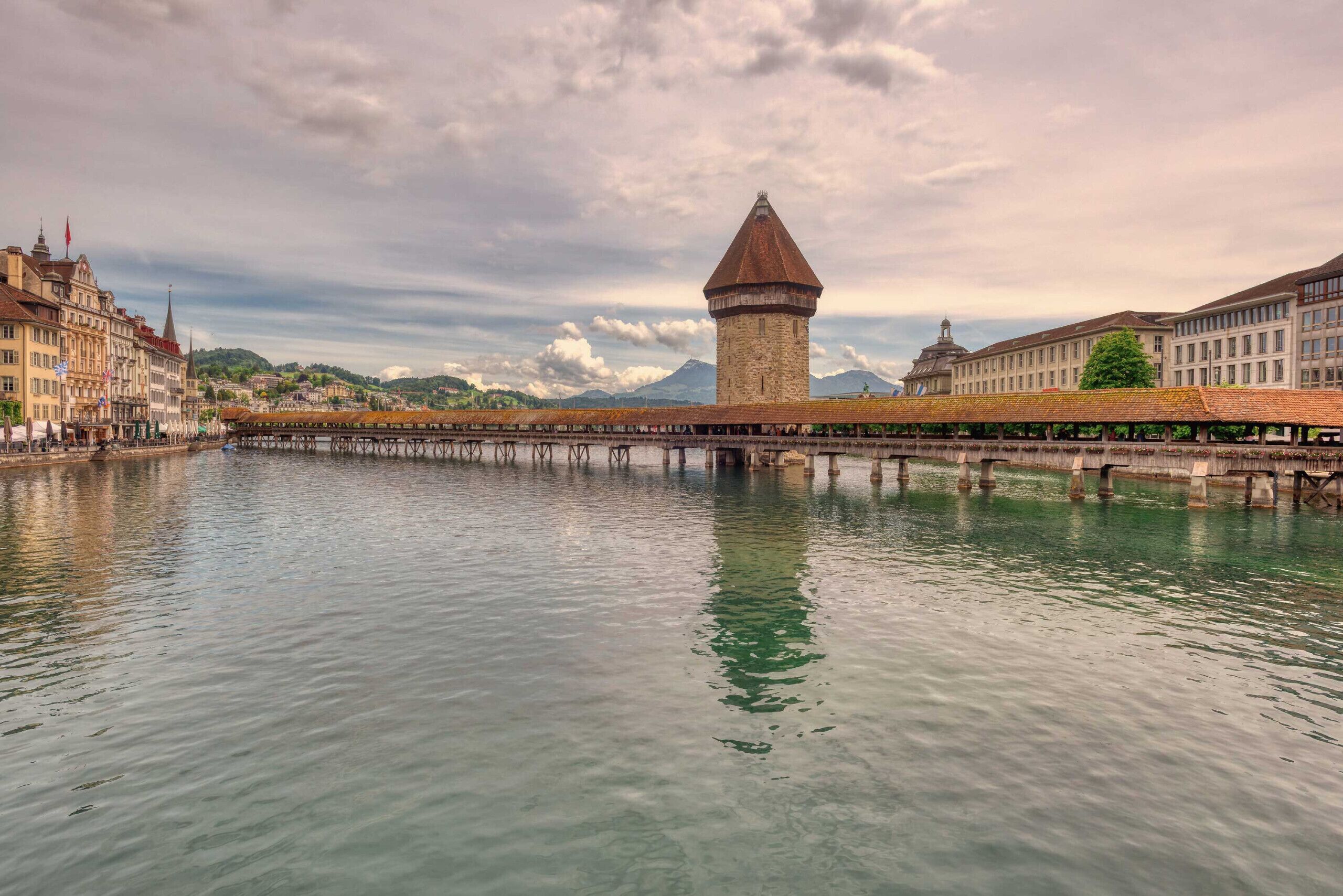 A historic tower by a calm river with a bridge, surrounded by buildings.
