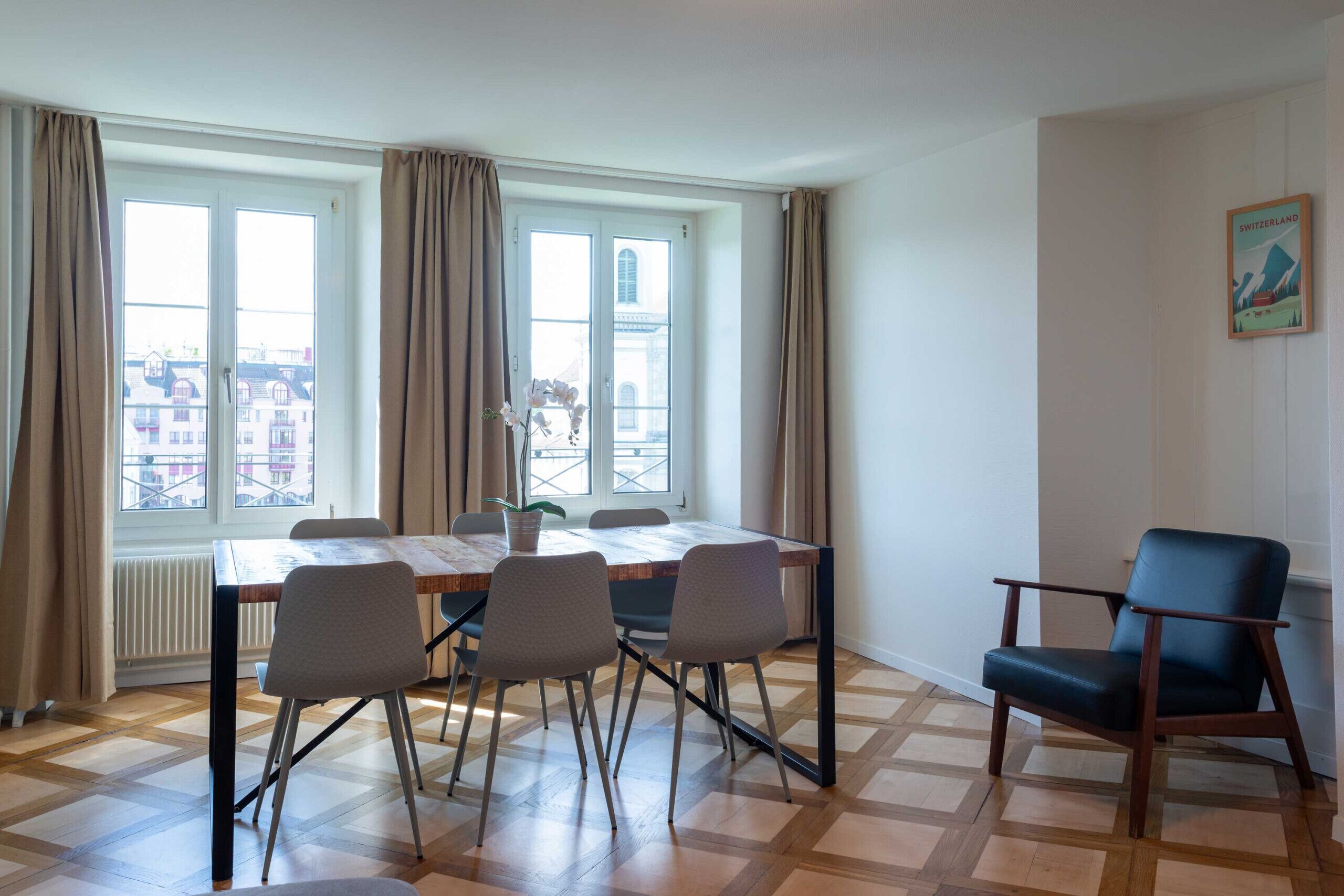 A room with a dining table, chairs, a potted plant, a window with curtains, and a framed picture on the wall.