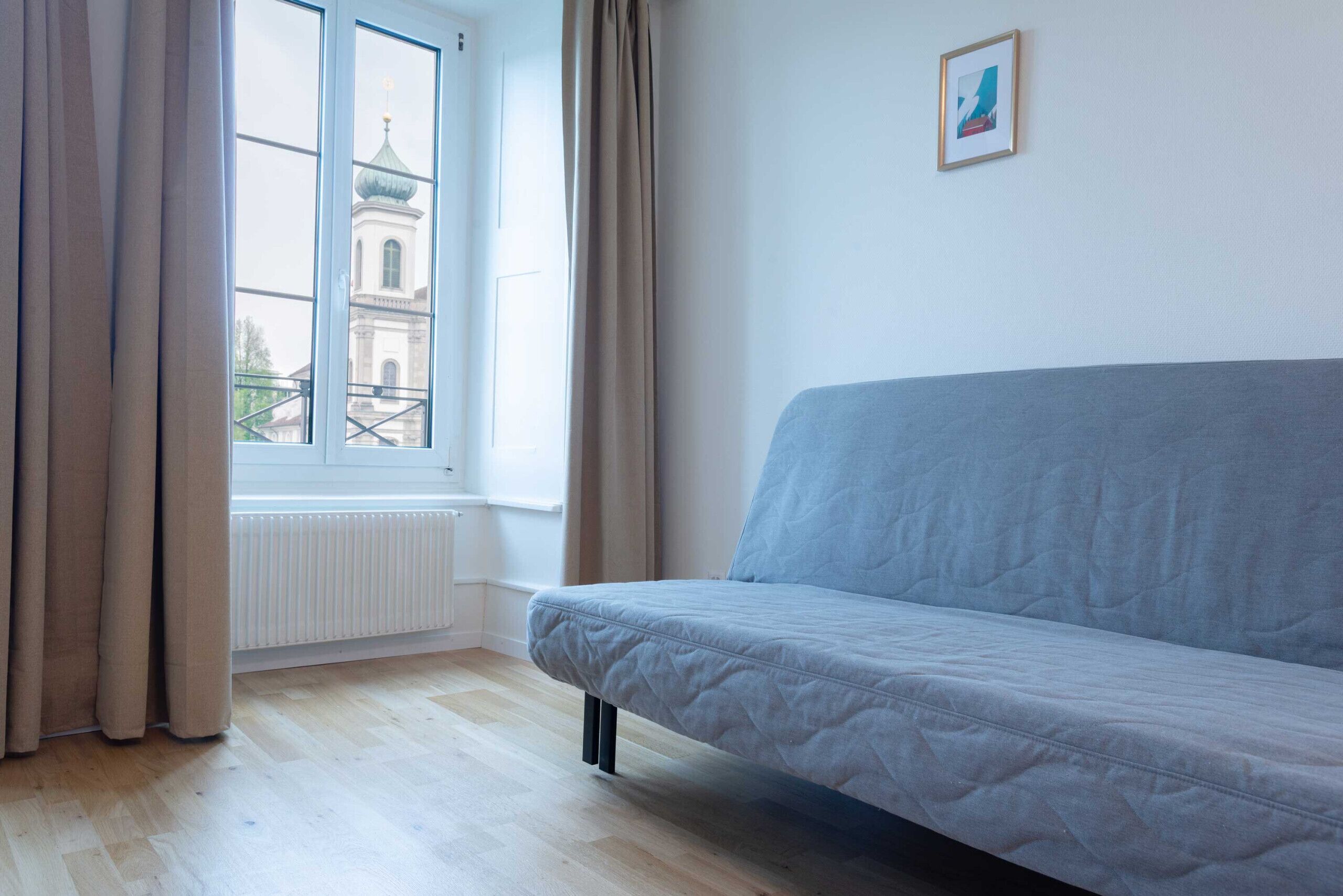 Een kamer met een lichtblauwe bank, een raam met gordijnen, een radiator en een ingelijst schilderij aan de muur.
