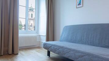 Een kamer met een lichtblauwe bank, een raam met gordijnen, een radiator en een ingelijst schilderij aan de muur.