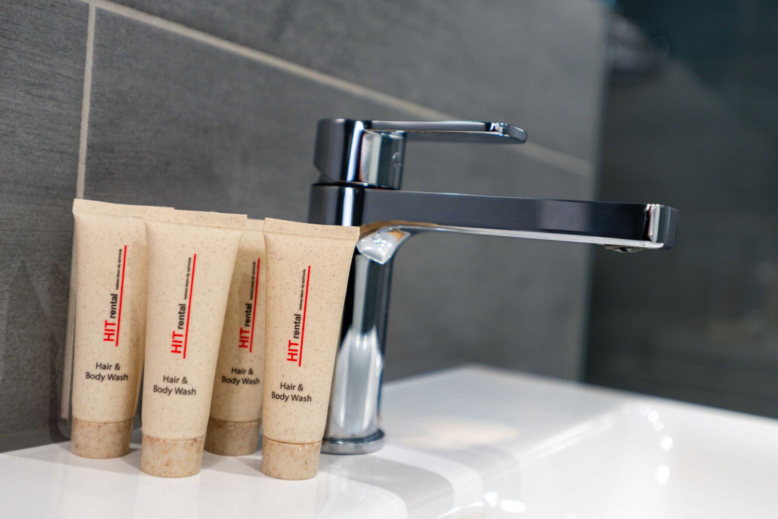 Une salle de bain avec un robinet chromé et quatre tubes de gel lavant pour les cheveux et le corps, de la marque HIT, mis à disposition pour les clients.