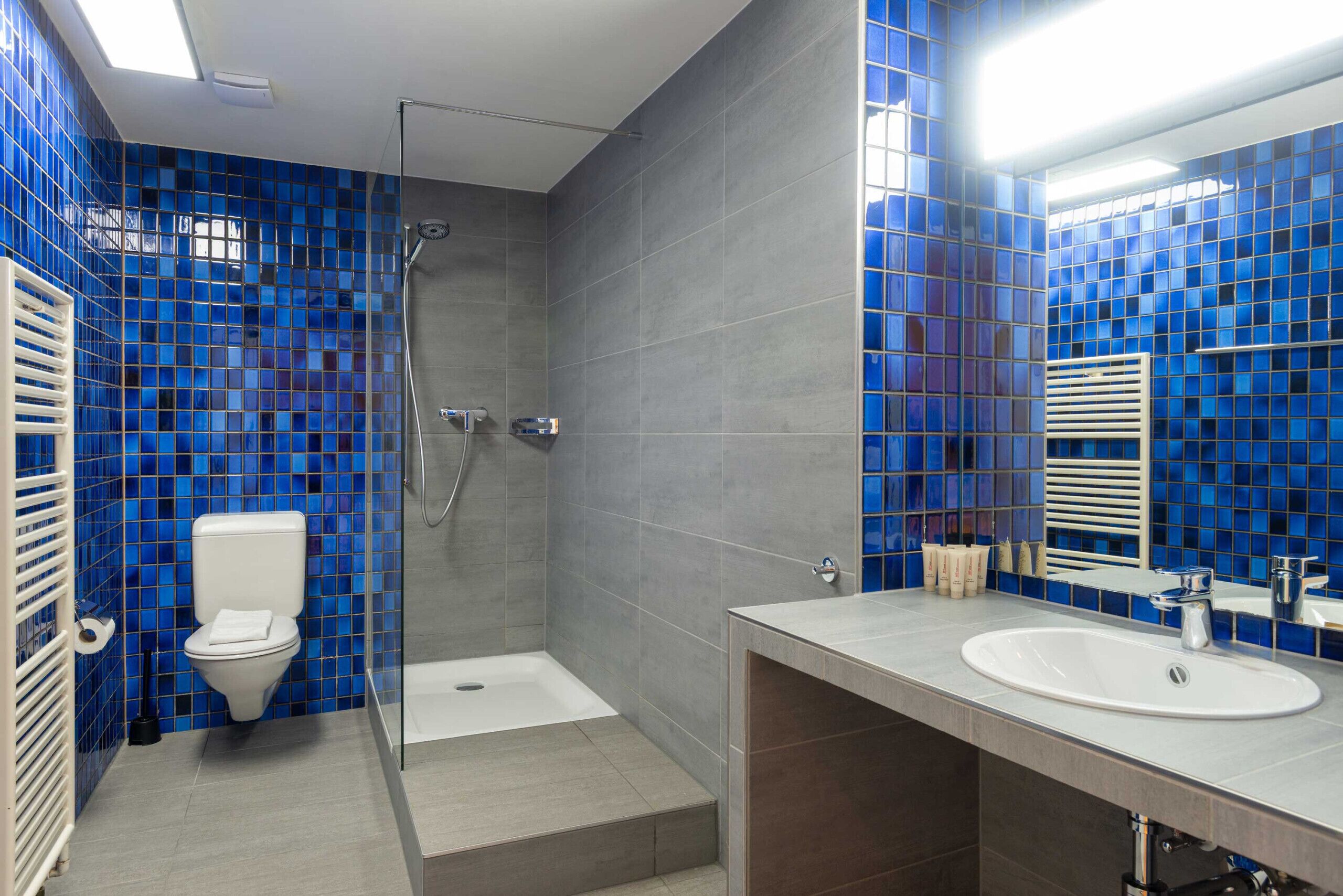 A bathroom with blue mosaic tiles, a white toilet, a glass shower enclosure, and a sink with a mirror.
