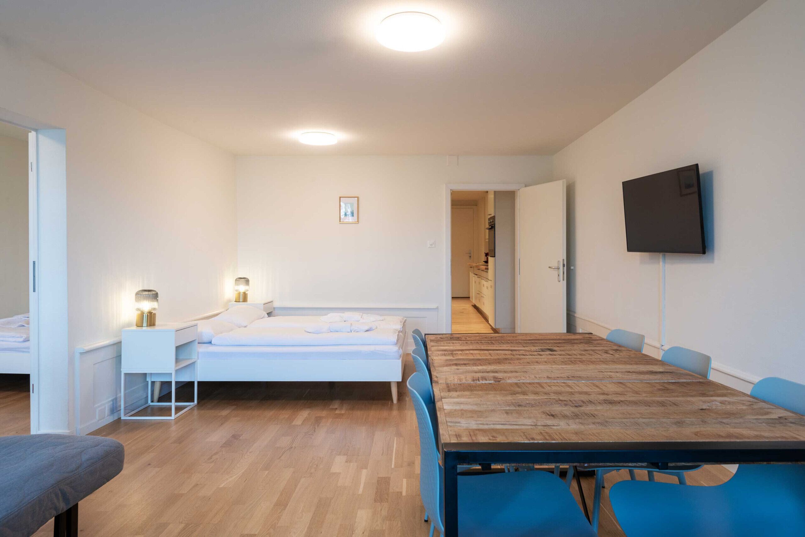 Una habitación moderna con una mesa de comedor de madera, sillas azules, una cama blanca y un televisor montado en la pared.