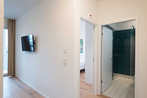 A hallway with a television mounted on the wall, a door leading to a bathroom with dark tiles, and a visible bed in the adjacent room.