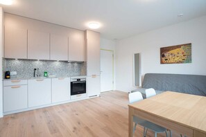 A modern kitchen with white cabinets, a granite countertop, and a built-in oven. There is a dining table with chairs and a gray sofa in the adjacent living area.