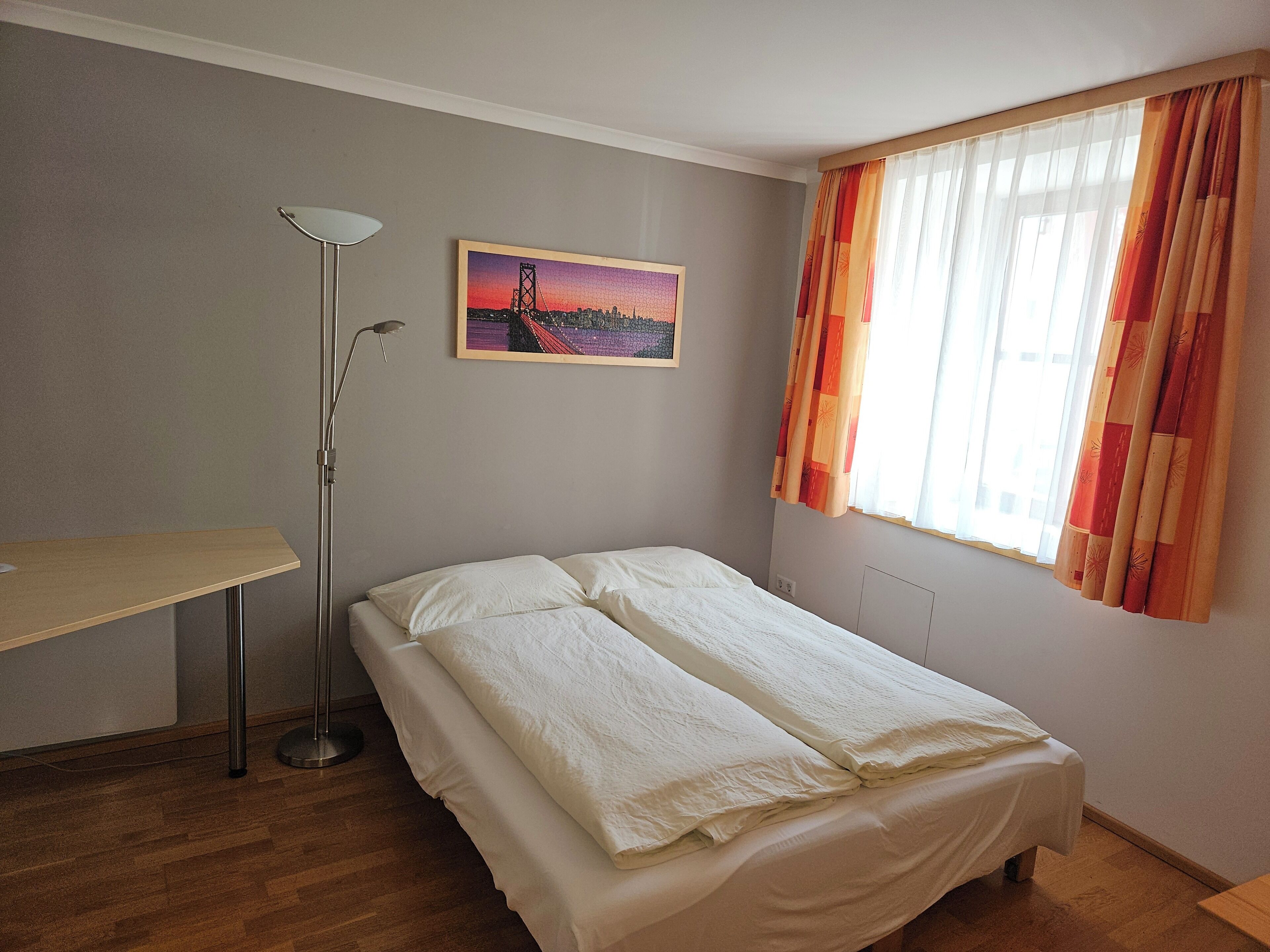 Et soveværelse med seng, skrivebord, gulvlampe og et vindue med gardiner.