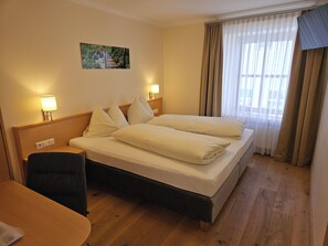 A hotel room with a bed, two bedside lamps, a small table, a chair, and a window with curtains.