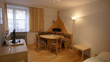 Una habitación compacta con suelo de madera, una mesa redonda con sillas, un banco, una mesita, un radiador, una lámpara y una cama.
