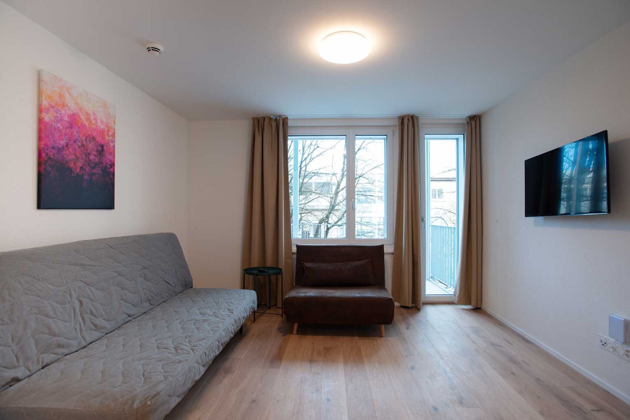 A modern living room with a grey sofa, a brown armchair, a small table, and a large window with curtains.