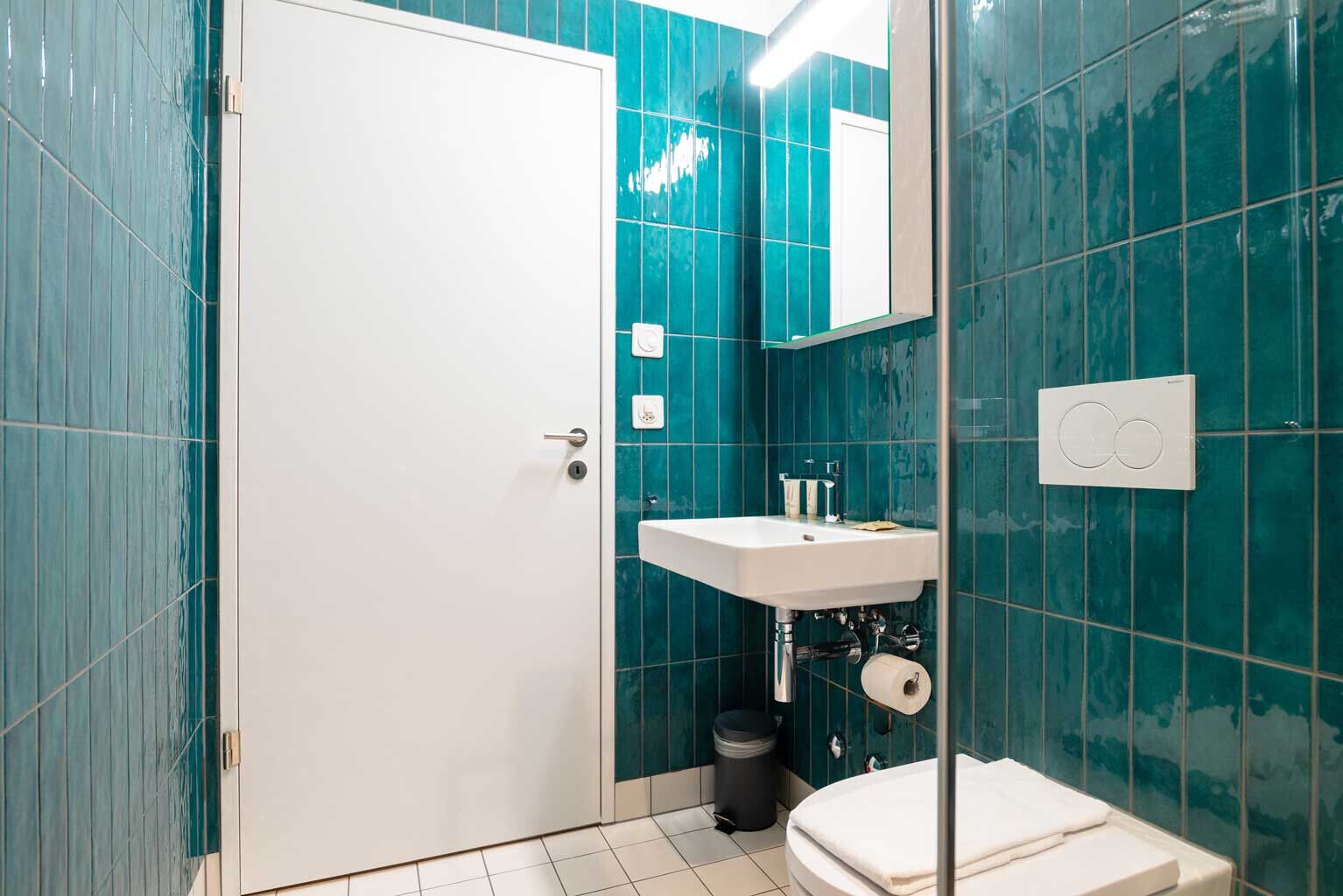 Ein Badezimmer mit türkisblauen Fliesen, einem weißen Waschbecken, einem Spiegel und einer Toilette.