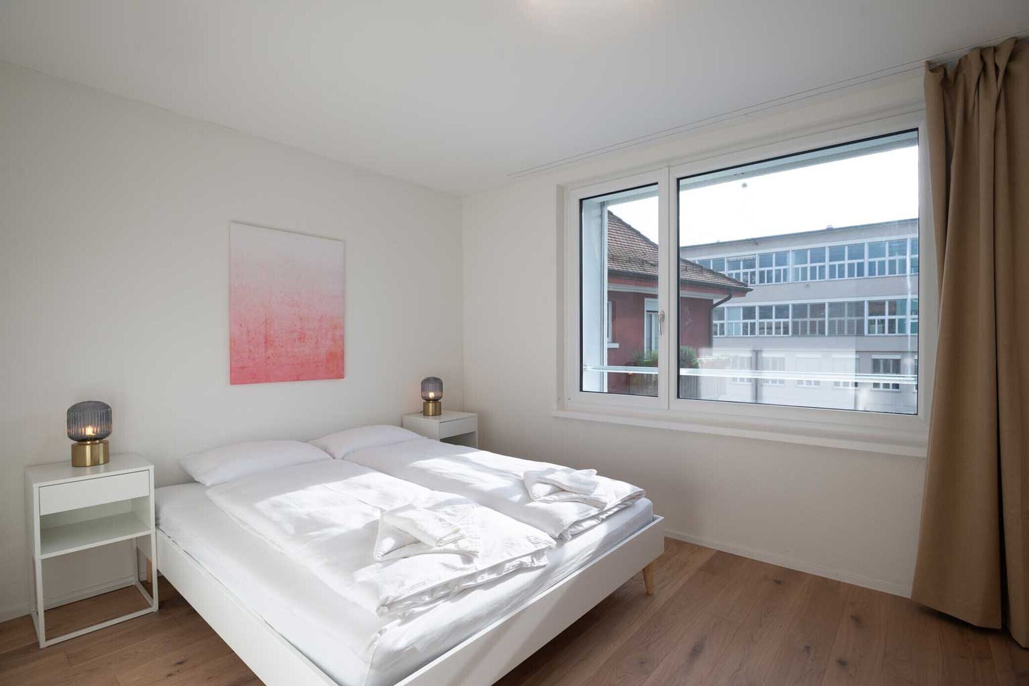 Een slaapkamer met een bed, nachtkastjes, een raam met gordijnen en uitzicht op gebouwen.