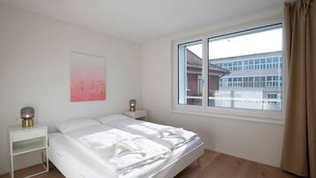Een slaapkamer met een bed, nachtkastjes, een raam met gordijnen en uitzicht op gebouwen.