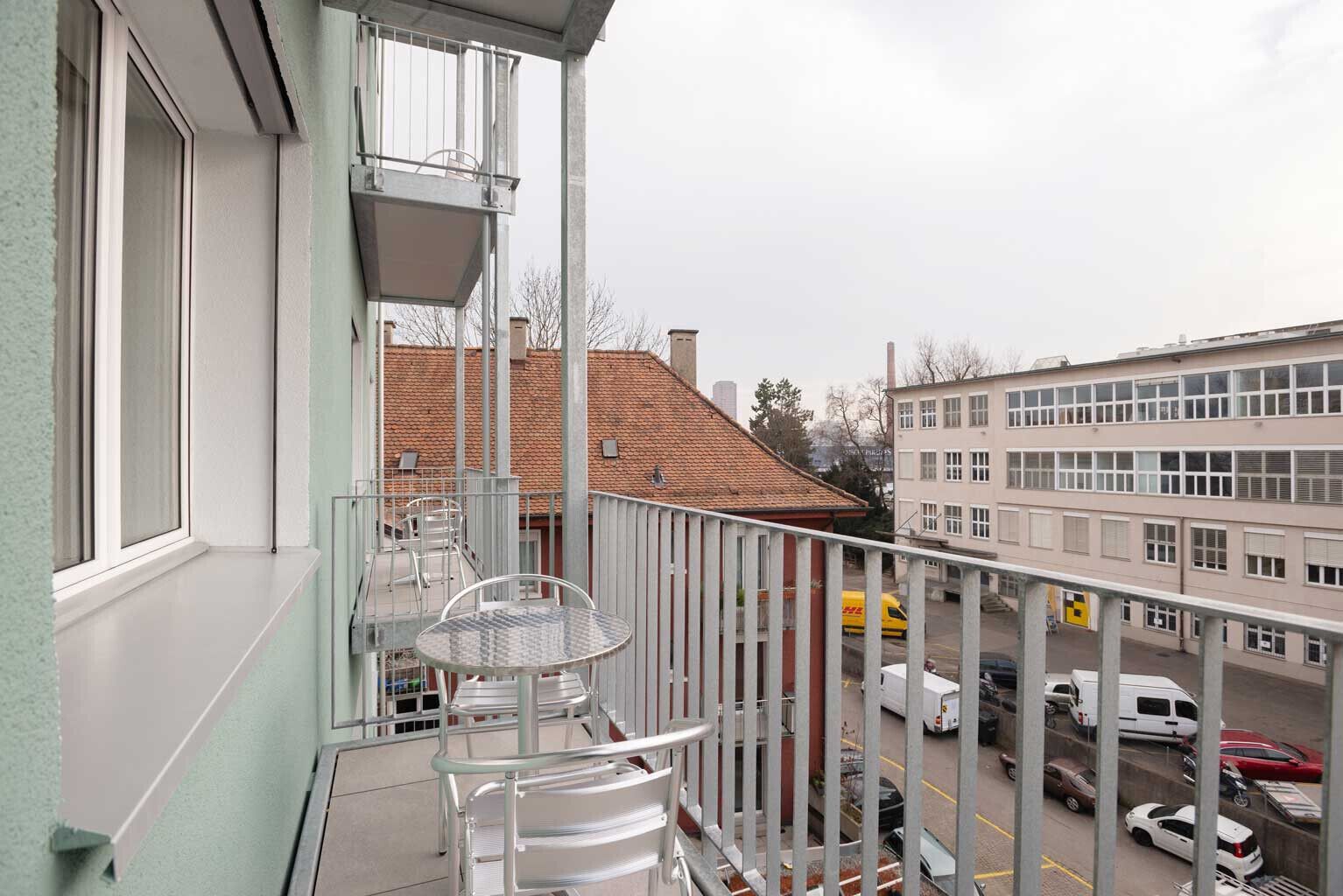A balcony with a table and chairs, overlooking a street with parked cars and buildings.