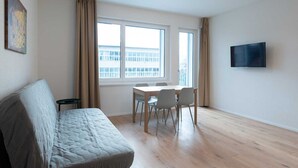 Un salon moderne comprenant un canapé, une table à manger avec des chaises, un téléviseur fixé au mur et de grandes fenêtres avec des rideaux.