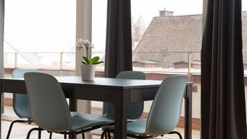 A dining area with a table and chairs, a potted plant, and a view of buildings through large windows.