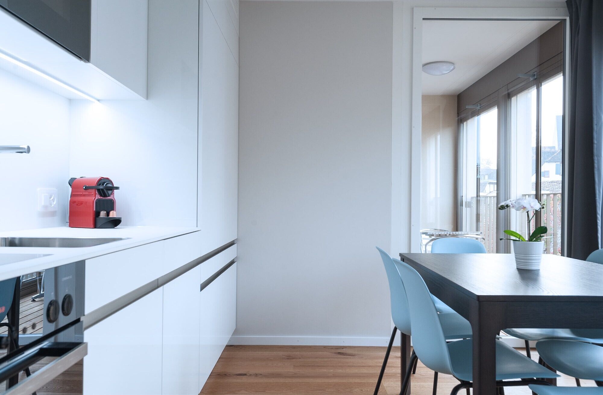 Una cocina moderna con armarios blancos, una cafetera roja y un comedor con mesa y sillas.